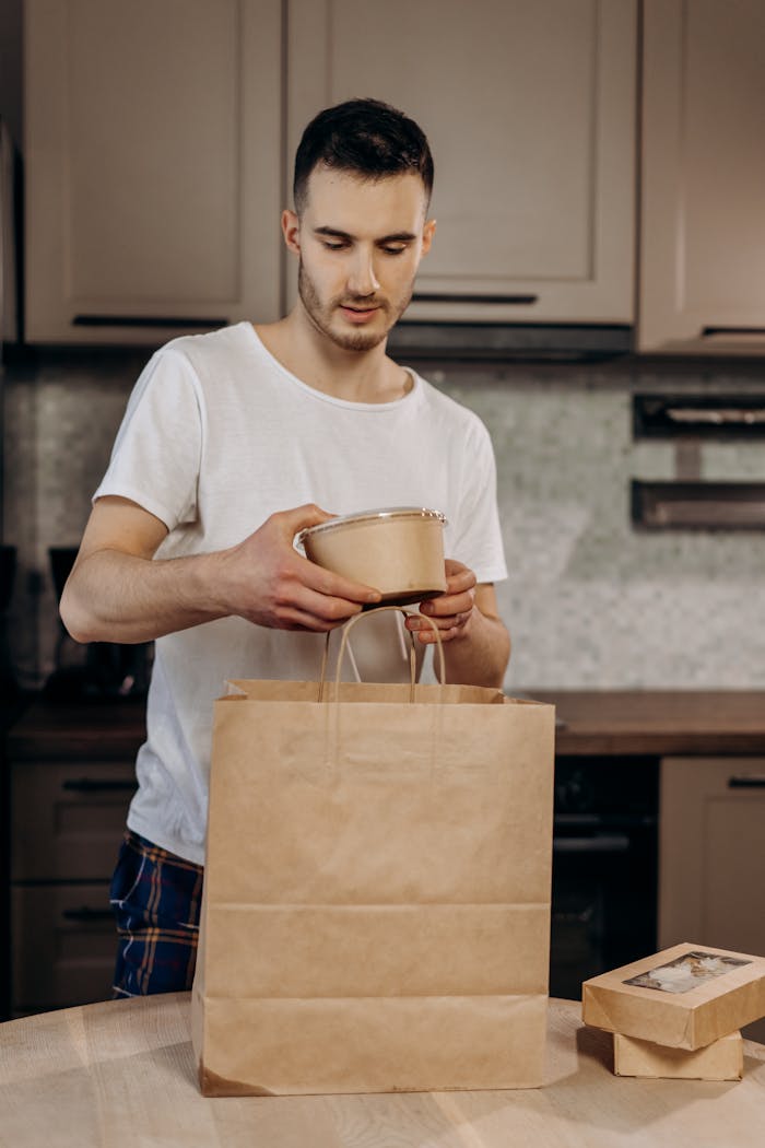 A young man in a kitchen packing takeout food into a brown paper bag, showcasing eco-friendly packaging.